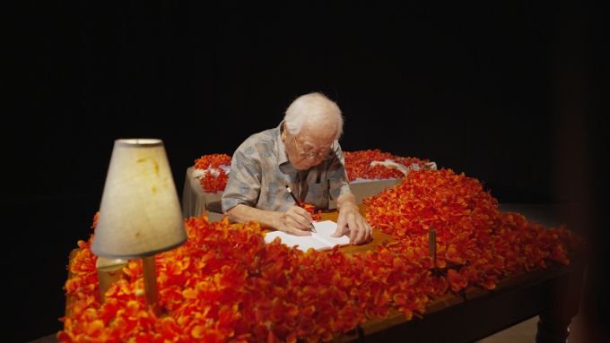 ベラウ（現地の言葉でパラオのこと）の花に囲まれた山城達雄さん。米国施政権下で受けた差別をきっかけに、人権のために闘う決意をし、沖縄人民党に入党、機関紙の記者として活動した。50代から小説を書きはじめ、『ベラウの花』などを著した。山城知佳子《Recalling(s)》2025年 ©Chikako Yamashiro. Courtesy of the artist